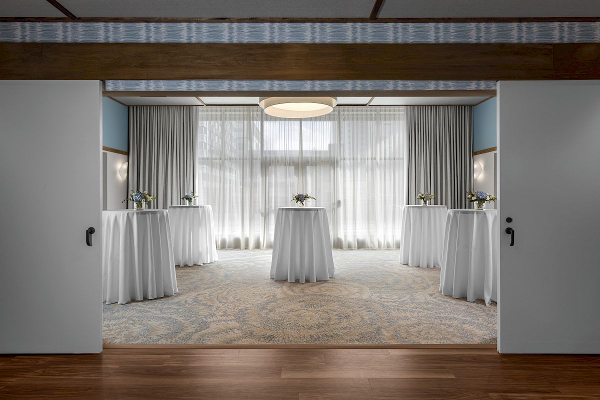 The image shows an elegantly decorated room, presumably for an event, with round tables covered in white cloths, floral centerpieces, and sheer curtains.