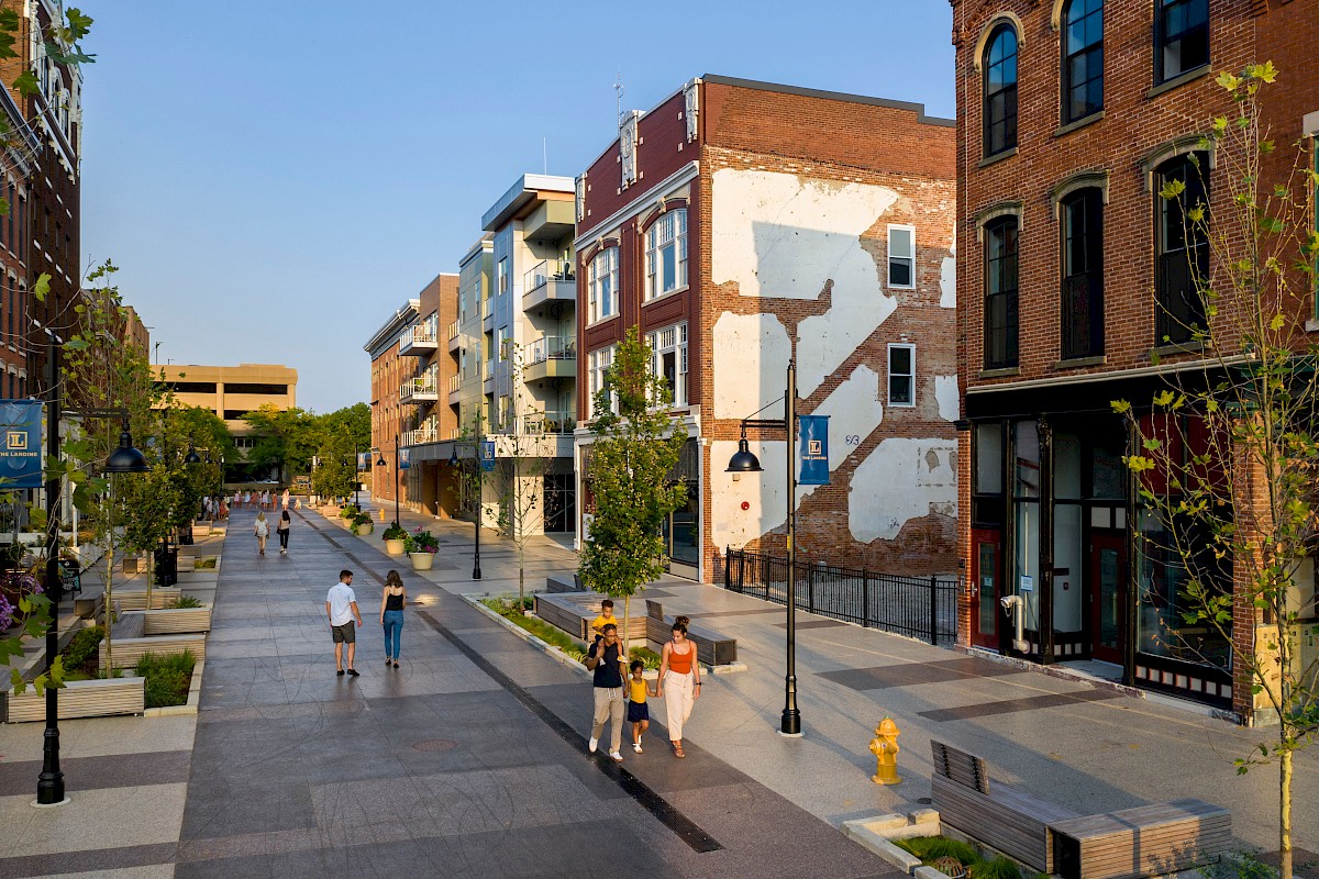 People walking along a pedestrian street lined with buildings, trees, and outdoor seating areas in an urban setting.