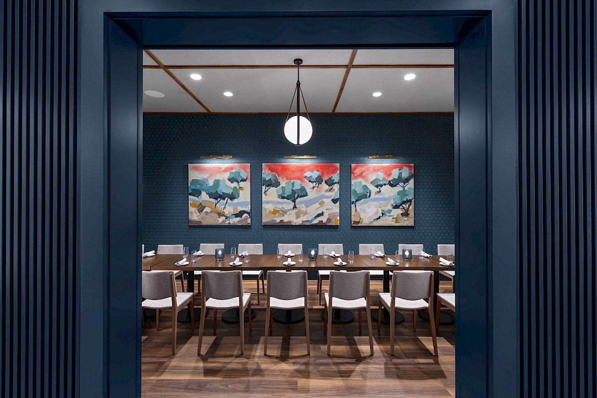 The image shows a modern dining room with a long table, white chairs, pendant light, and three abstract paintings on a dark blue wall.