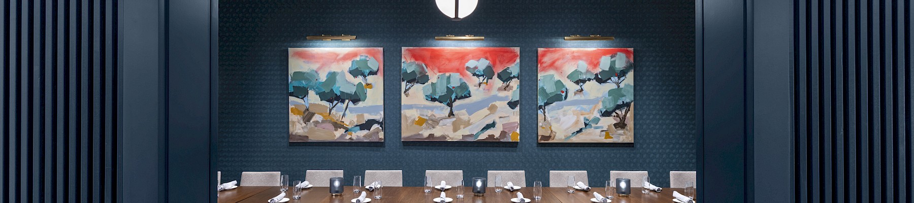 The image shows a dining room with a long table surrounded by chairs, three abstract paintings on the wall, and a round ceiling light.