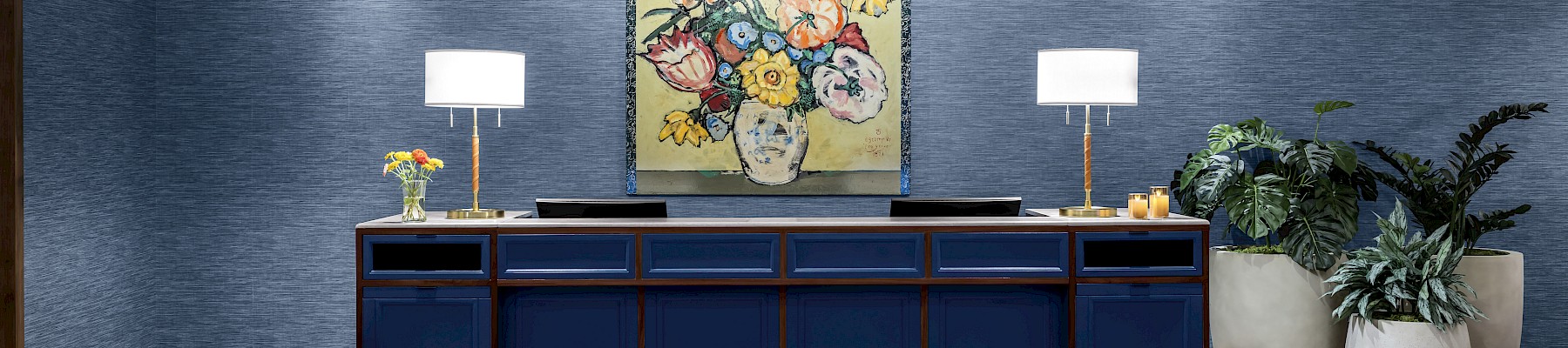 A room with blue walls, a sideboard, table lamps, a colorful painting, potted plants, and an orange cushioned seat on wooden flooring.