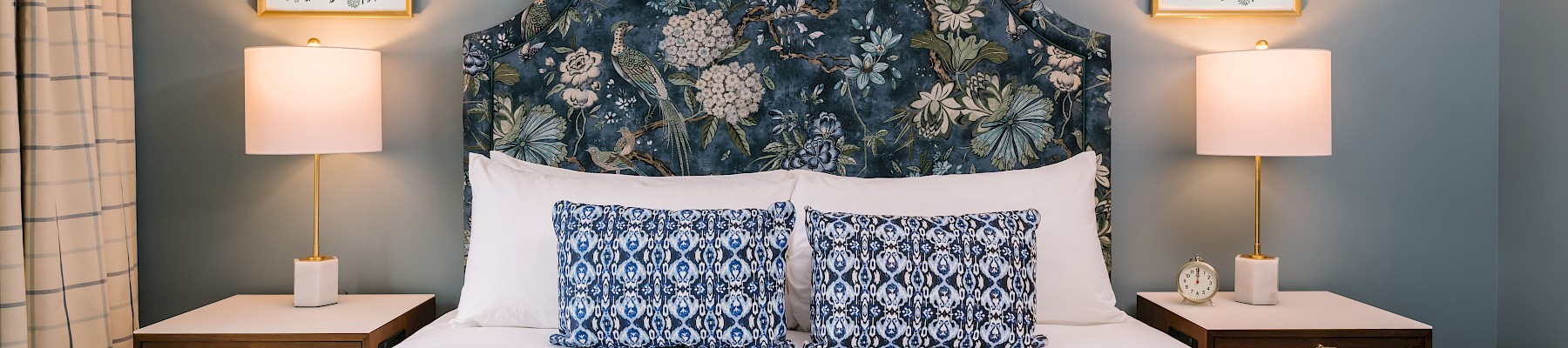 A stylish bedroom with a floral headboard, patterned pillows, two lamps on nightstands, and framed botanical art on the wall.