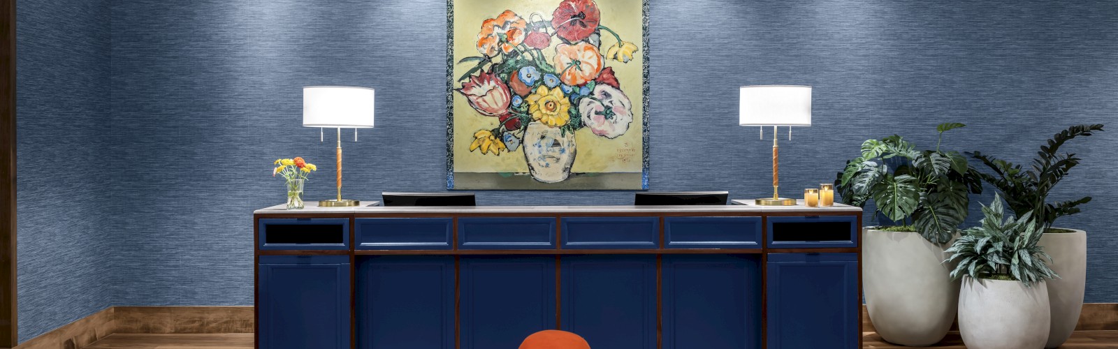 A modern interior with a blue cabinet, two white lamps, a floral painting, plants in pots, and an orange sofa on wooden flooring.