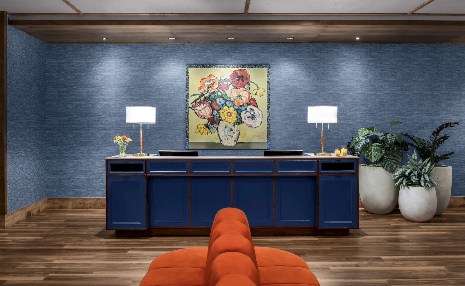 A waiting area features a blue cabinet with lamps, a floral painting, potted plants, and an orange seat on a wood floor.