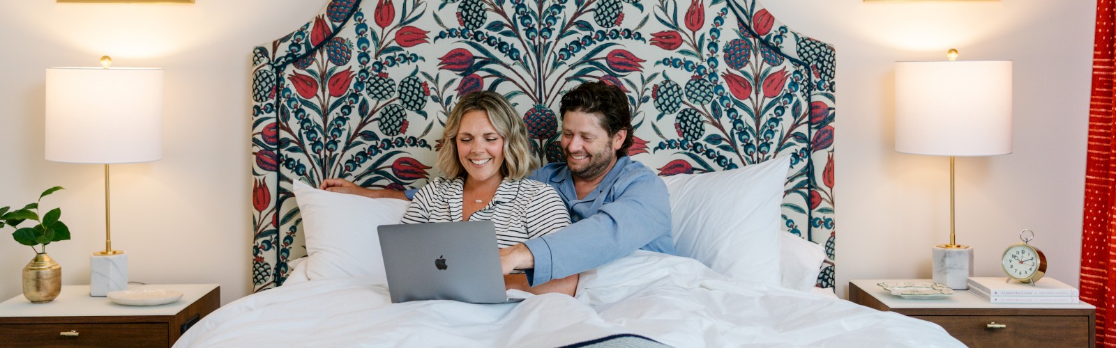 A couple is sitting in bed, sharing a laptop. The headboard has a floral pattern, with framed botanical prints and lamps on either side.