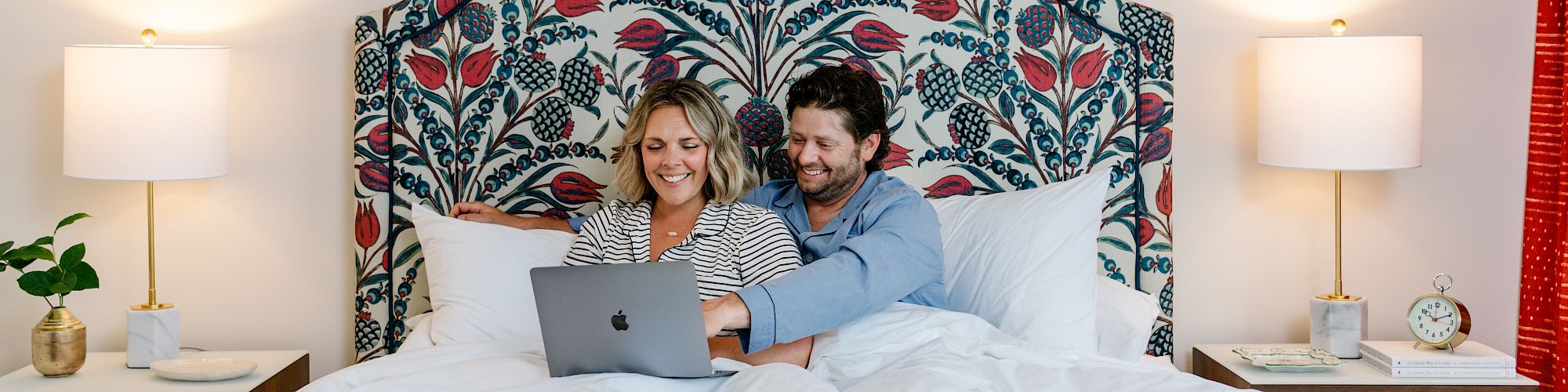 A couple is sitting in bed, sharing a laptop. The headboard has a floral pattern, with framed botanical prints and lamps on either side.