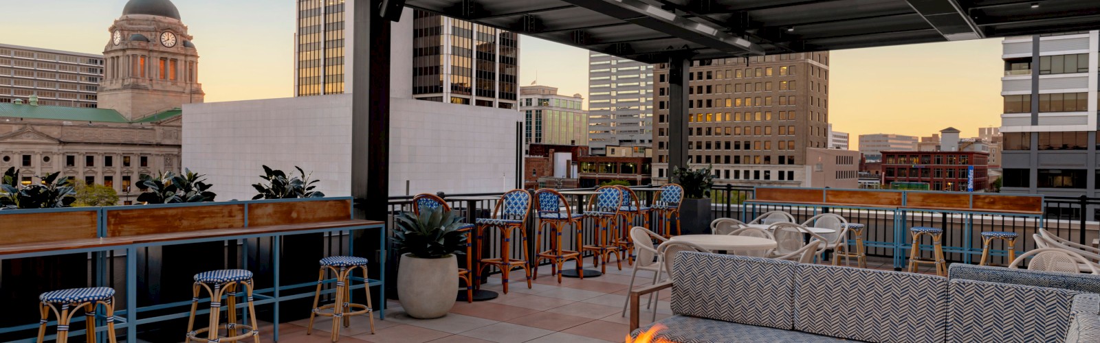 A rooftop patio with a fire pit, cushioned seating, bar stools, and city skyline views, including a domed building in the background.