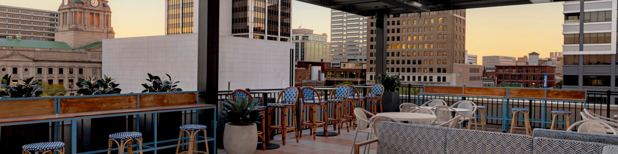 A rooftop patio with a fire pit, cushioned seating, bar stools, and city skyline views, including a domed building in the background.