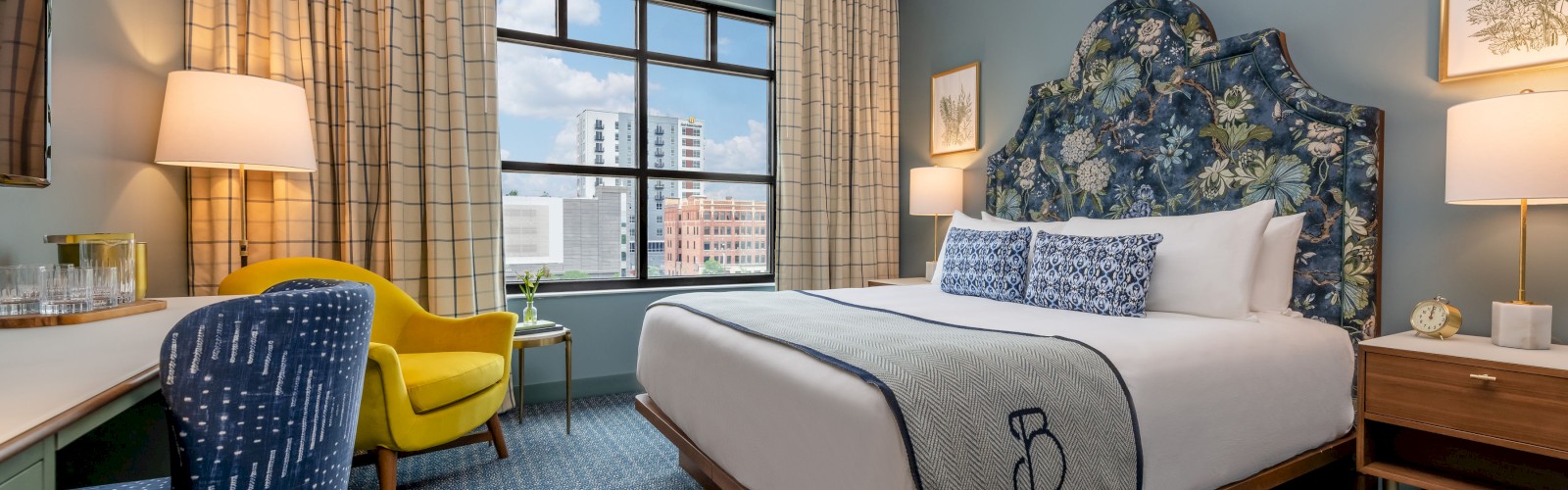 A stylish hotel room features a bed, two chairs, desk, lamps, and framed art. A window offers a city view.