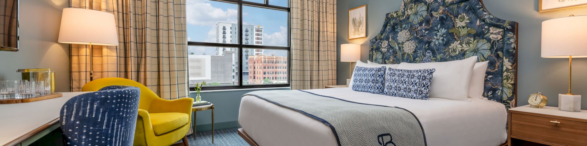 A stylish hotel room features a bed, two chairs, desk, lamps, and framed art. A window offers a city view.