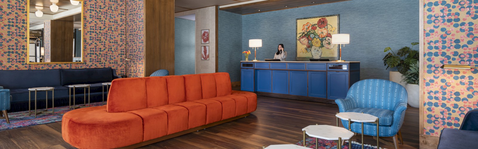 The image shows a stylish lobby with colorful furniture, including a long orange sofa, blue armchairs, small tables, and a reception desk.