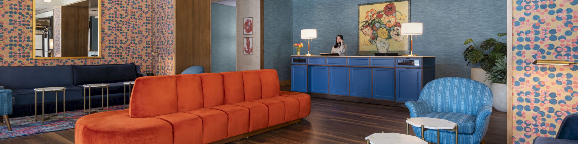 The image shows a stylish lobby with colorful furniture, including a long orange sofa, blue armchairs, small tables, and a reception desk.