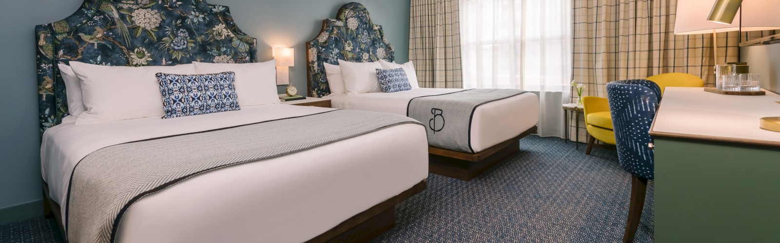 The image shows a hotel room with two double beds, colorful headboards, a desk with a lamp and chair, and a large window with curtains.