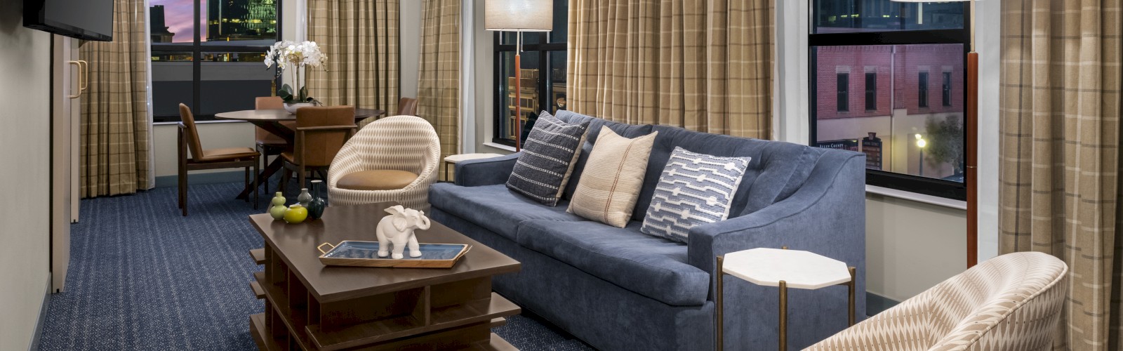 A modern living room with a blue sofa, two chairs, a coffee table, floor lamps, a TV, and large windows with city views at sunset.
