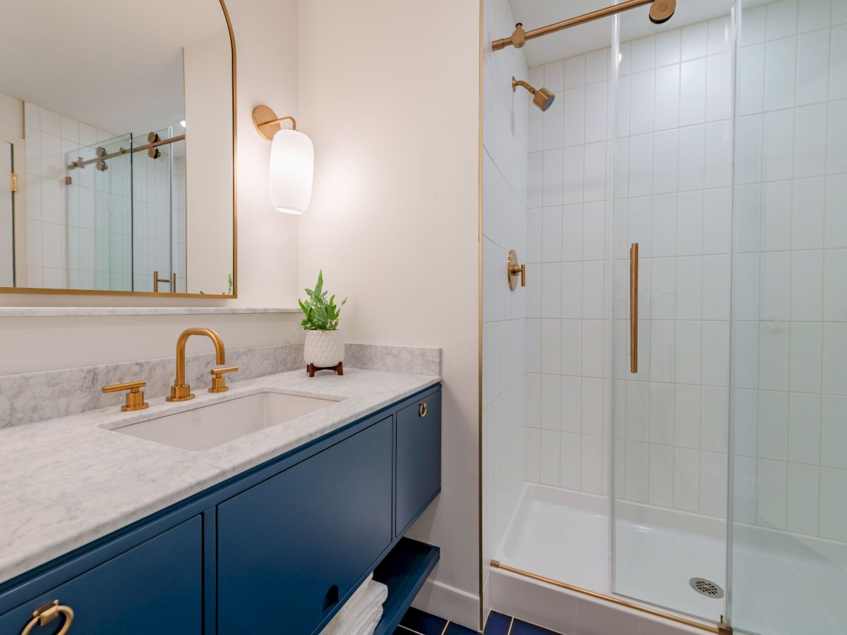 A modern bathroom features a blue vanity with a marble countertop, gold fixtures, wall lamp, mirror, and a glass shower door with gold hardware.