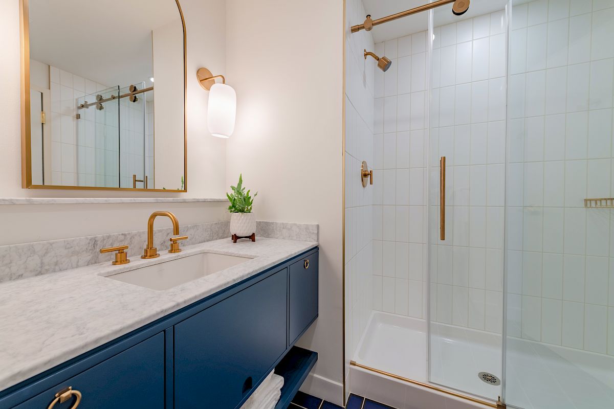 A modern bathroom with a blue vanity, marble countertop, gold fixtures, a mirror, a small potted plant, and a glass shower enclosure.