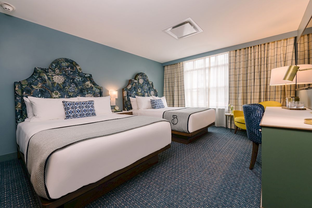 The image shows a hotel room with two double beds, patterned headboards, a desk, chairs, a lamp, and a large window with curtains.