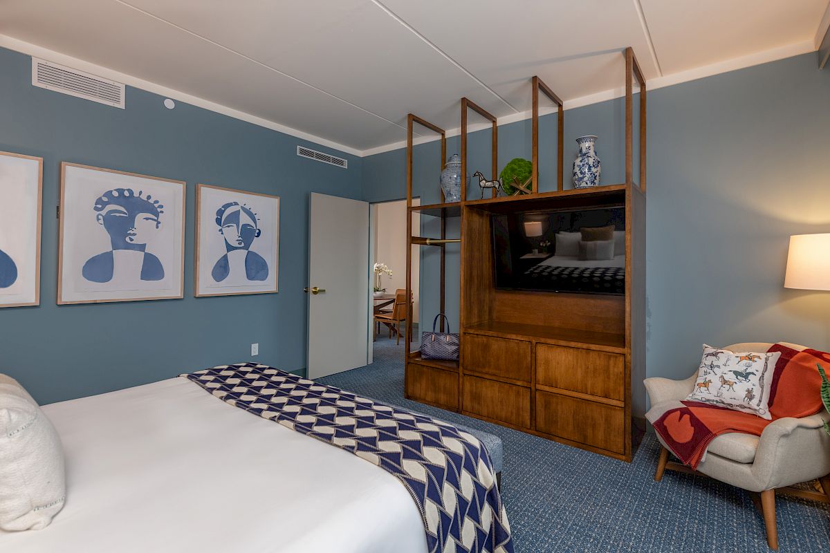 A cozy hotel room features a bed, modern artwork, a TV shelf, an armchair with a pillow, and a side lamp on a blue-themed decor.