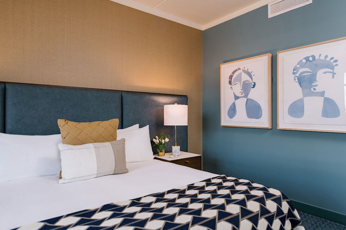 A neatly made bed with decorative pillows, a bedside table with a lamp and vase, and two framed abstract artworks on the wall complete the decor.