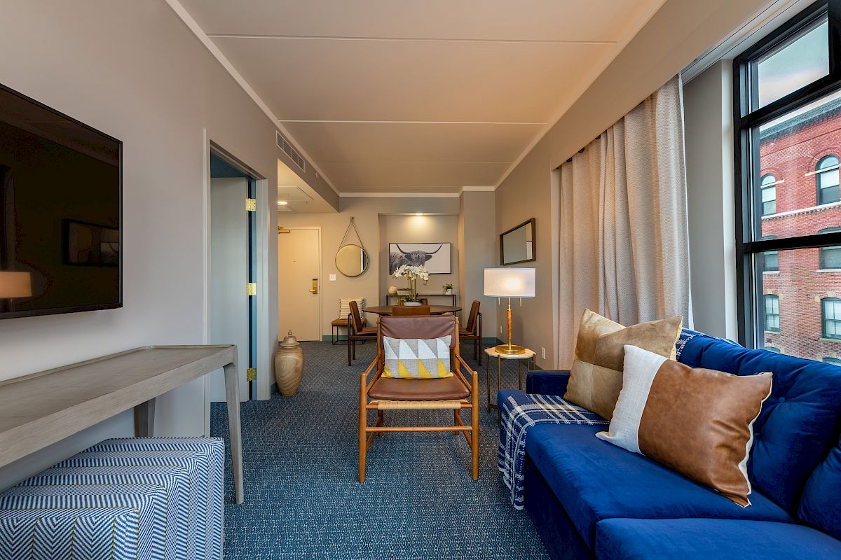 This image shows a modern, cozy living room with a blue sofa, wooden chairs, a TV, and decorative pillows. A large window provides natural light.
