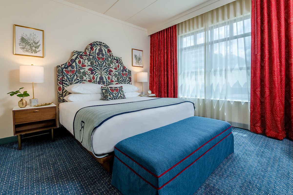 The image shows a well-decorated hotel room with a large bed, blue bench, bedside tables, lamps, red curtains, and framed artwork on the wall.