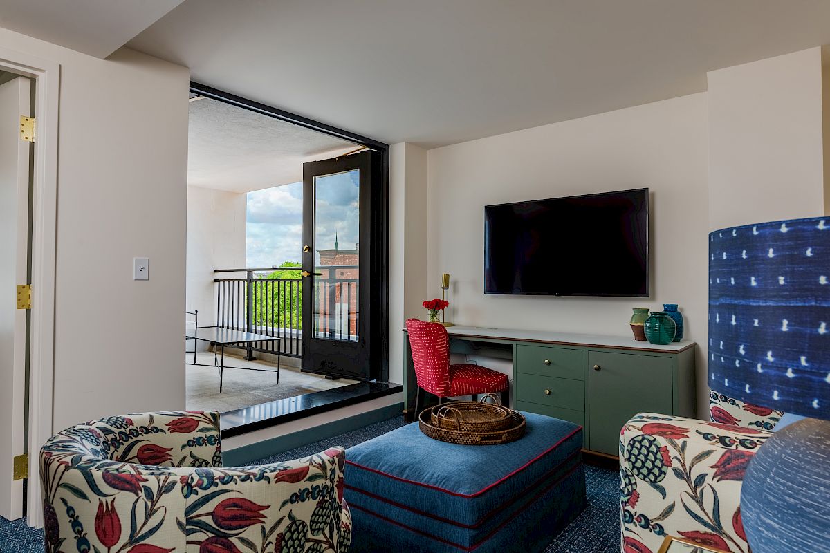 A cozy living room features a TV, two floral-patterned chairs, an ottoman, and a balcony with outdoor seating and a view.
