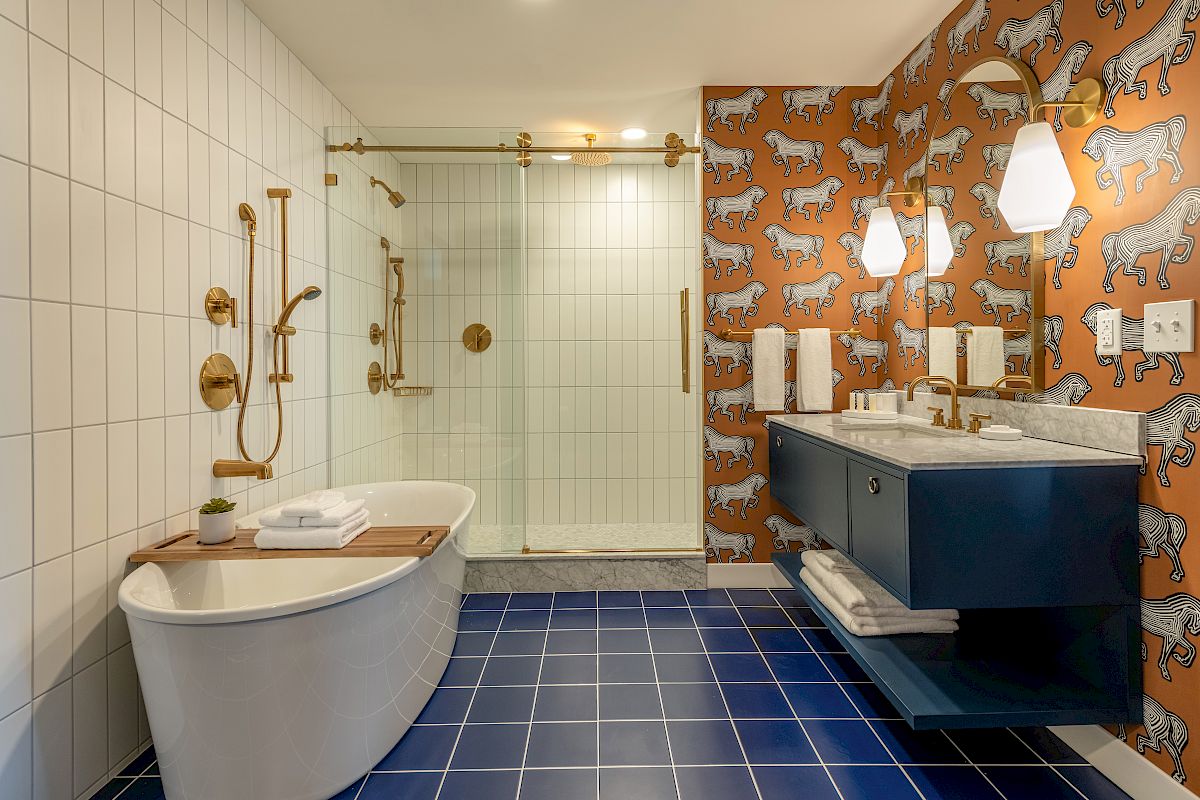 A modern bathroom with a freestanding tub, blue-tiled floor, glass shower, double vanity, and unique wallpaper with white animal illustrations.