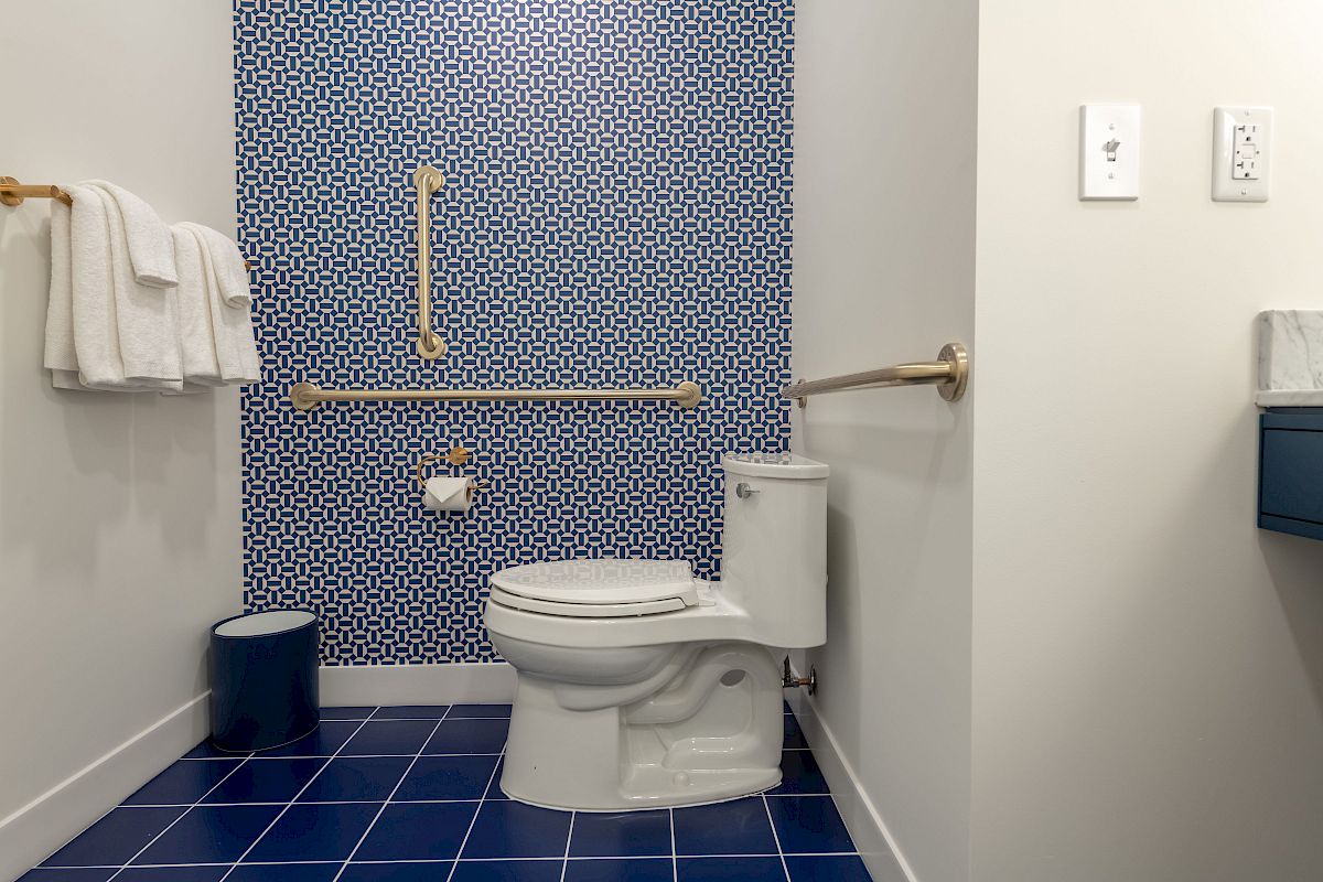 A bathroom with a toilet, blue tiled wall and floor, towel rack, small waste bin, grab bars, and light switches on the right wall, ending the sentence.