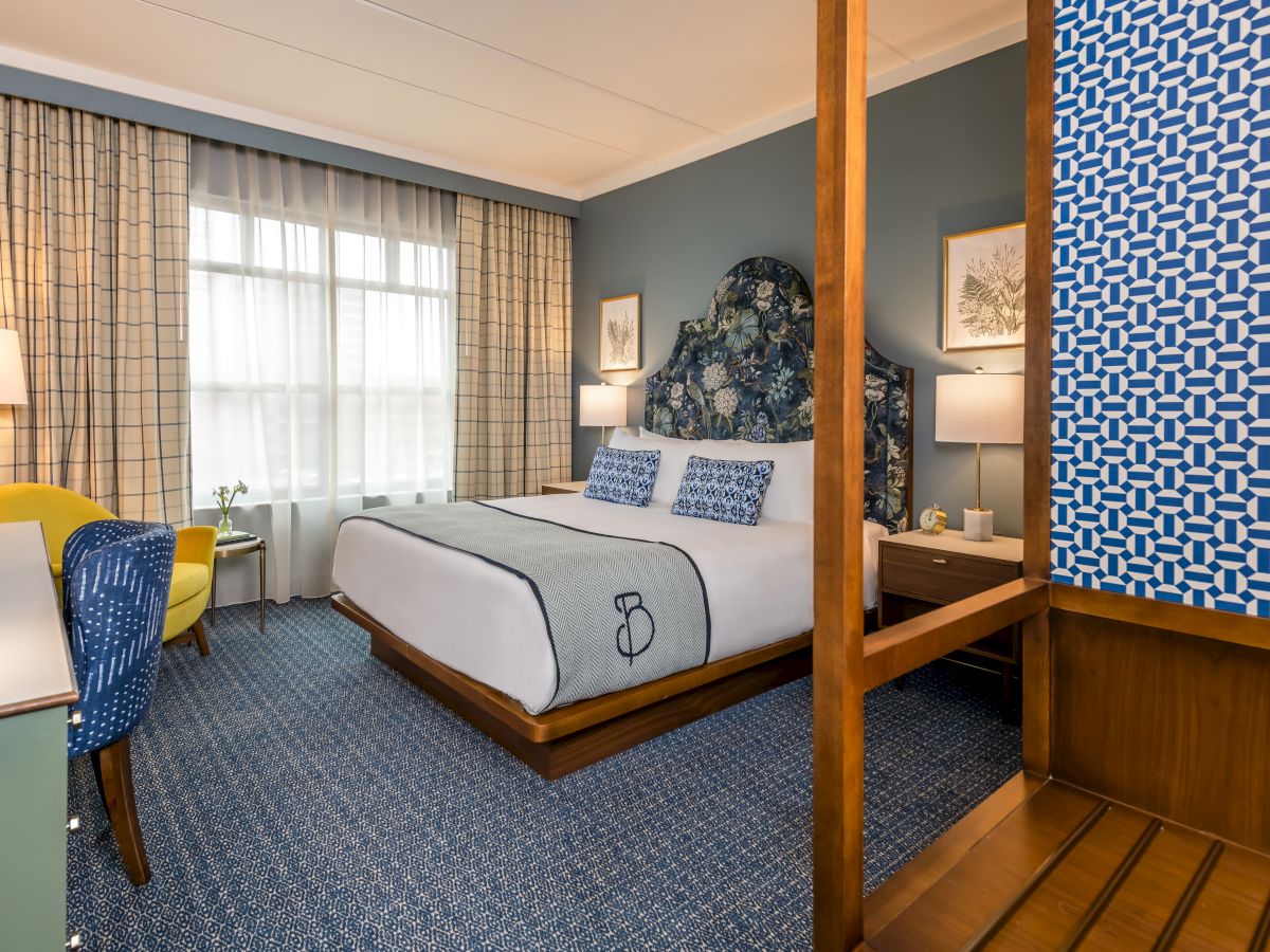 A neatly arranged hotel room features a king-size bed, patterned cushions, a blue accent wall, a desk with a chair, and a window with curtains.