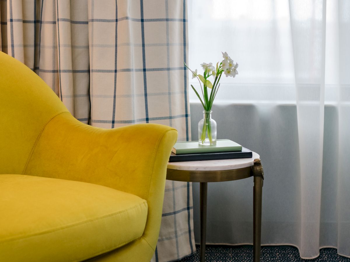 A yellow chair, a side table with a flower vase, a book, and checked curtains in front of a window with sheer white curtains.