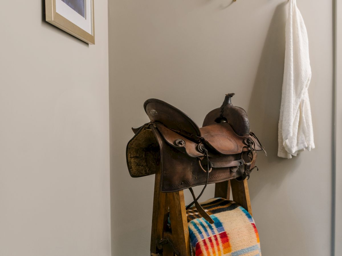 A saddle with a colorful blanket draped over it stands in a corner. A framed picture and a coat hook with a white garment are on the wall.