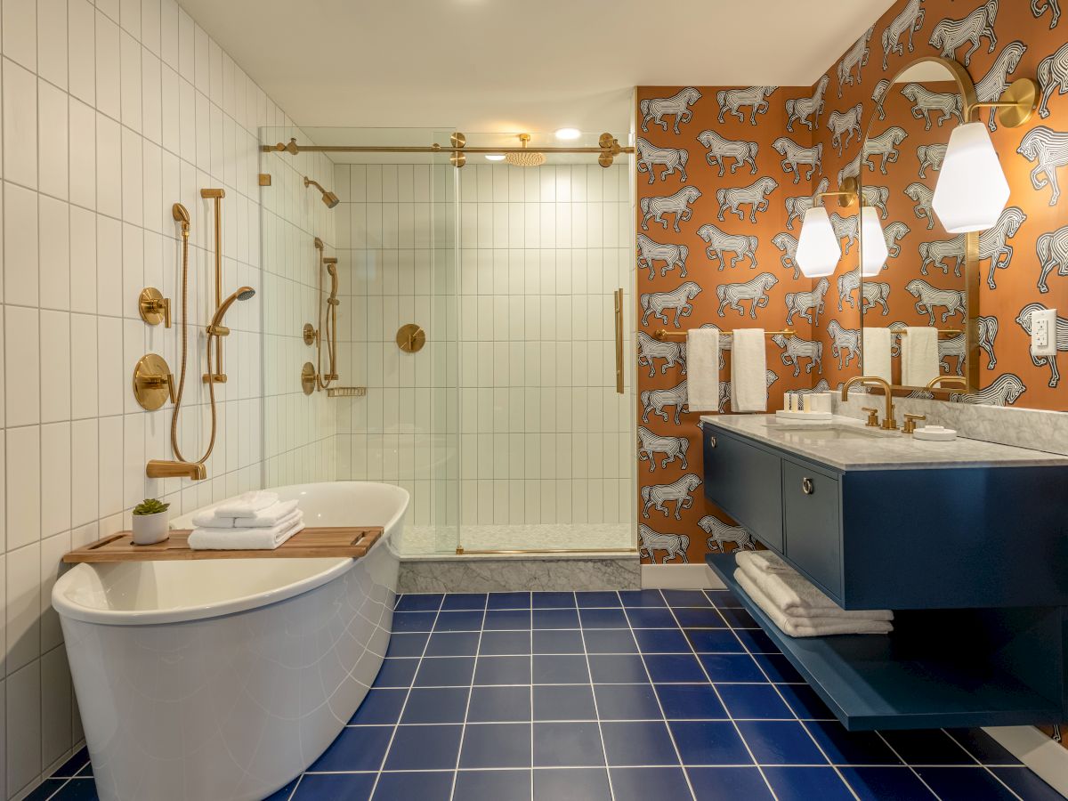 This image shows a modern bathroom with white tiles, a freestanding tub, glass shower, blue vanity, leopard-patterned wallpaper, and blue floor tiles.