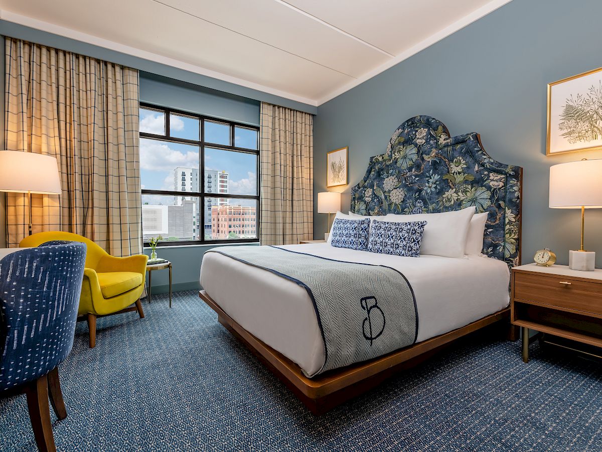 A modern hotel room features a bed with patterned headboard, desk with chair, yellow armchair, bedside tables, and large window with city view.