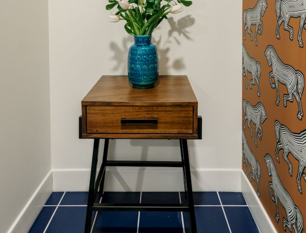 A small wooden table with a blue vase and green plant, framed artwork above, blue tiled floor, and orange wallpaper with zebra pattern on one wall.