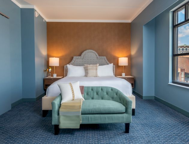 A neatly arranged hotel room features a king-size bed with a cushioned headboard, two bedside tables with lamps, and a small green sofa.