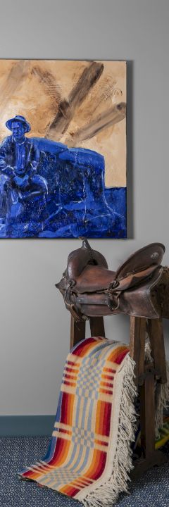 The image shows a corner with a blue-toned painting, a brown saddle on a stand, a colorful blanket, and two decorative hooks on a gray wall.