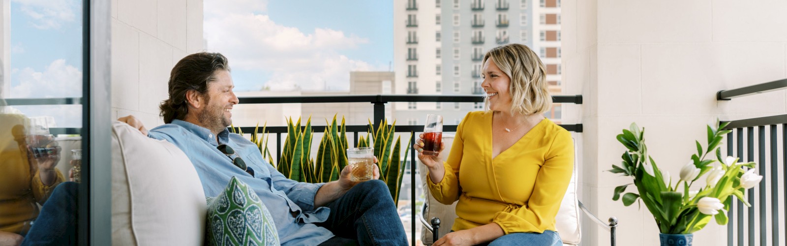 A man and a woman sit on a balcony sofa, enjoying drinks and conversation. A vase with flowers is on a table beside them, with a city view.