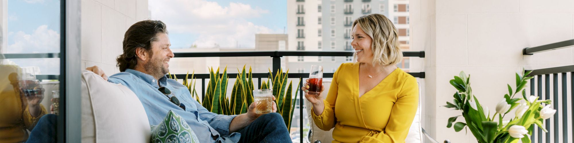 A man and a woman sit on a balcony sofa, enjoying drinks and conversation. A vase with flowers is on a table beside them, with a city view.