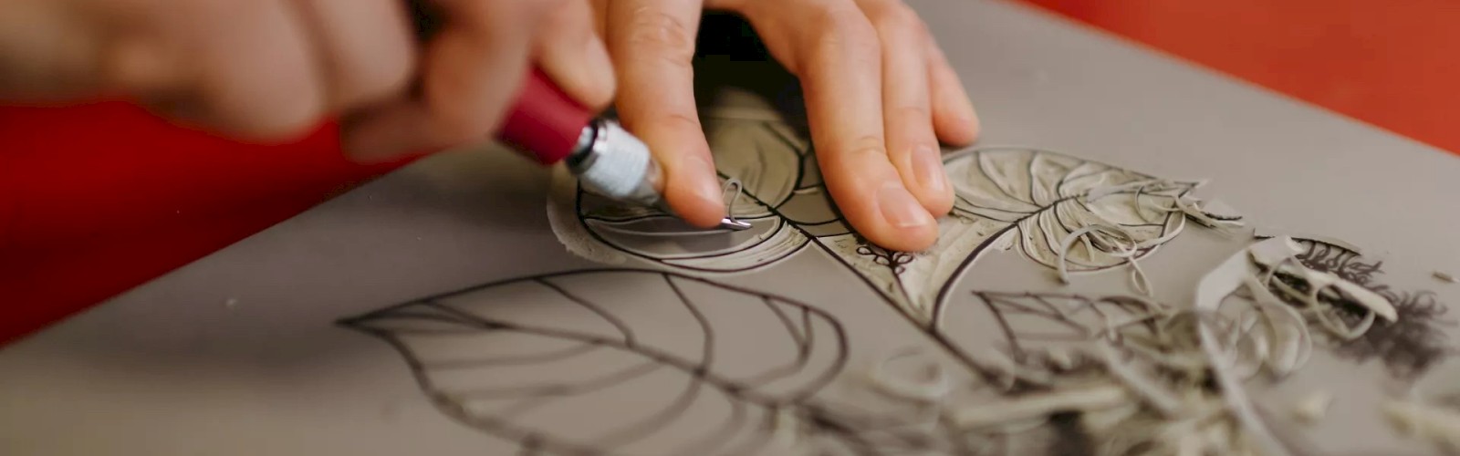 A person is using a carving tool to create an intricate design with leaves and flowers on a piece of material, possibly linoleum or wood.