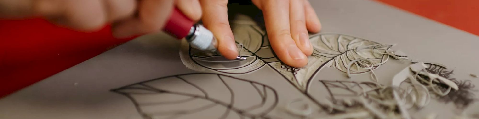 A person is using a carving tool to create an intricate design with leaves and flowers on a piece of material, possibly linoleum or wood.