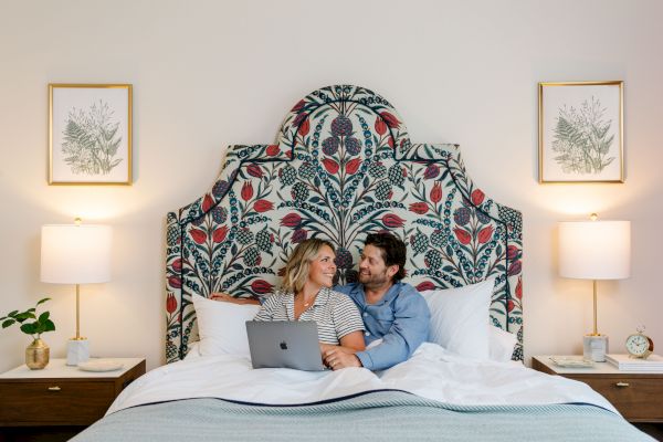 A couple is sitting on a bed with a colorful headboard, using a laptop, and smiling at each other. The room has matching lamps and decor.