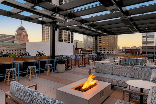 A rooftop lounge with a fire pit, cushioned seating, bar stools, and city buildings in the background at sunset.