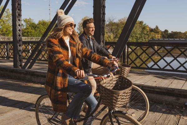 Two people are riding bicycles on a wooden bridge, enjoying the sunny weather, both smiling and dressed in casual, warm clothing.
