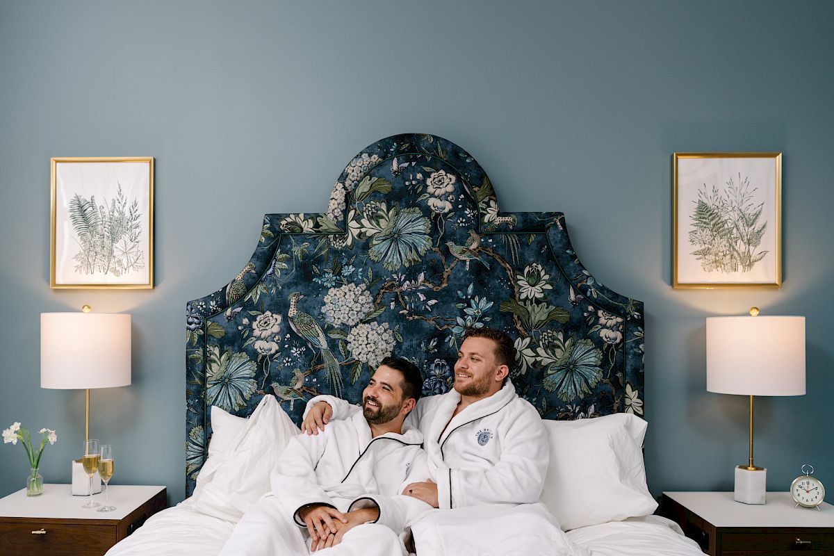 Two people wearing white robes sit on a bed against a floral headboard, flanked by lamps and framed art on the walls, in a cozy room.