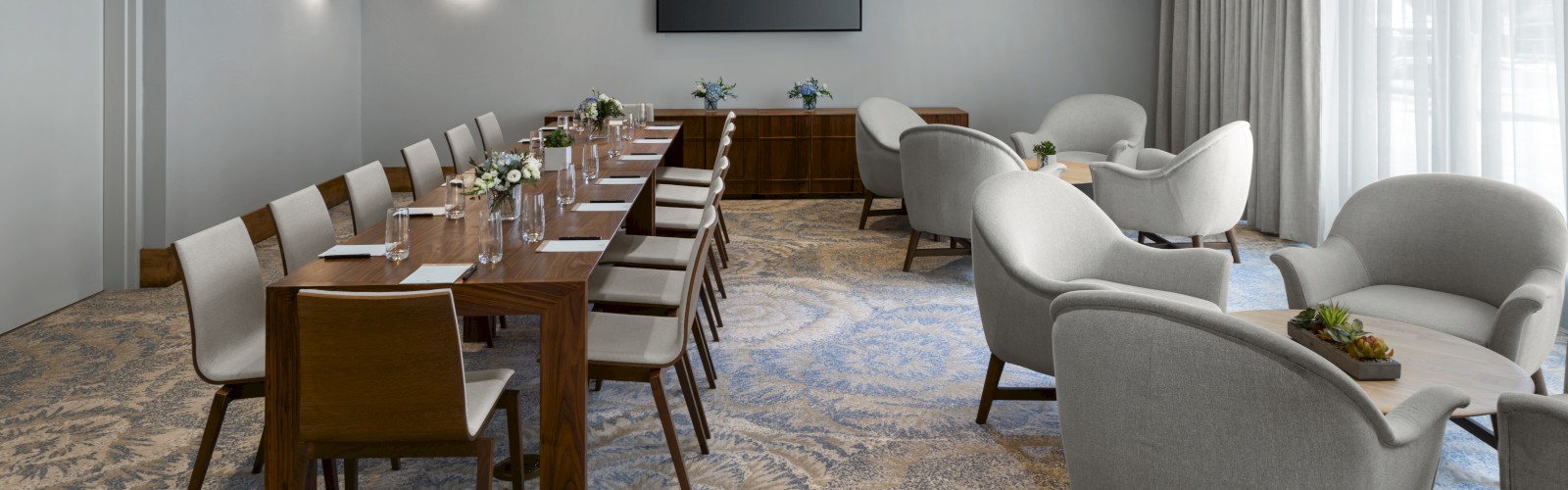 A modern meeting room with a long table, chairs, a TV, and grey armchairs arranged in a cozy setting near a window, ending the sentence.
