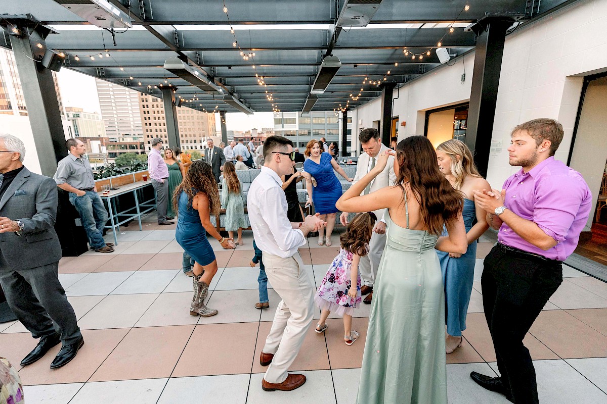 People are dancing and socializing on a rooftop terrace, surrounded by city buildings, under string lights.