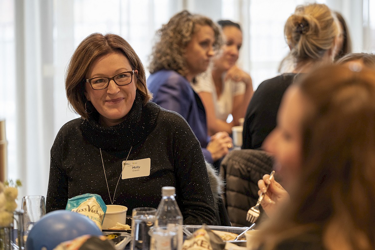 A woman with glasses, wearing a name tag, sits at a table with food and drinks, while others in the background are engaged in conversation.