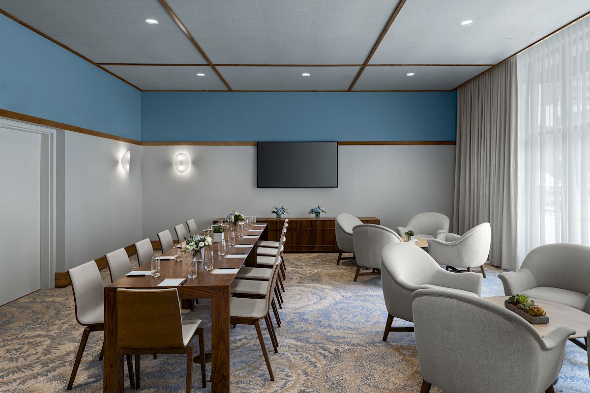 A modern conference room with a long table, chairs, a TV, armchairs, and a sideboard. The room has blue and white walls with a patterned carpet.
