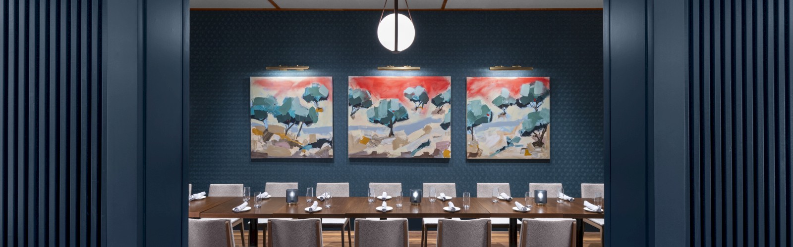 A modern dining room with a long table, surrounded by chairs, three colorful paintings, and a pendant light overhead.