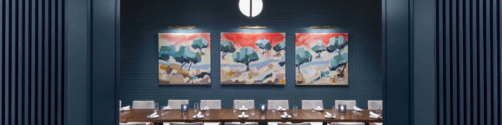 A modern dining room with a long table, surrounded by chairs, three colorful paintings, and a pendant light overhead.